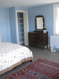 This large bedroom has a queen bed with two side tables and reading lamps, a dresser with mirror, a small closet, a writing table, easy chair, bookshelves, Turkish rugs, ceiling fan, and a bathroom. Its 3 windows face east (toward the church next door) and south (towards the front yard.) You can see the original wood floor, with wide boards.